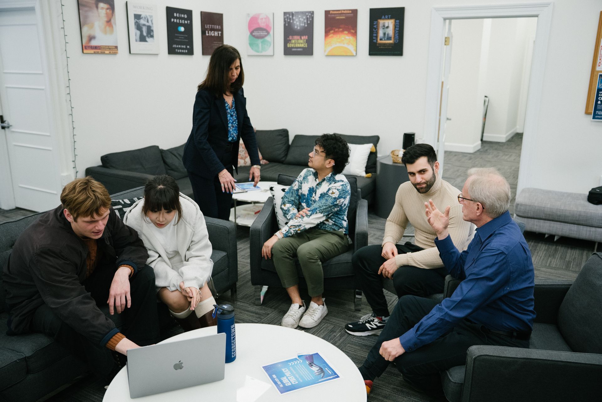 A group of people are gathered in a room and two students look at a laptop.