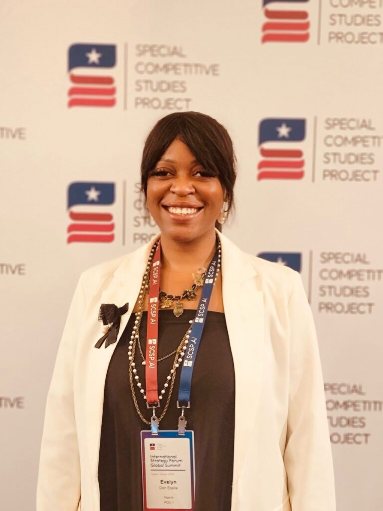 Evelyn Dan Epelle (CCT’20) smiles at the International Strategy Forum Global Summit. She is wearing a white blazer and name badge, standing in front of a backdrop with the logo of the Special Competitive Studies Project.