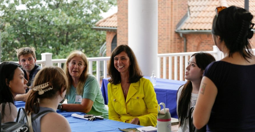 Dr. Jeanine Turner sits at a table surrounded by students and faculty. She is wearing a yellow dress and smiling at a student.